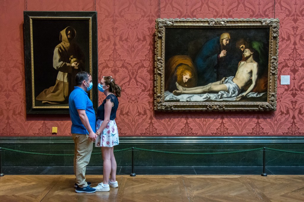 Man and woman in blue face mask in a museum holding hands facing each other. Classical Paintings in the background